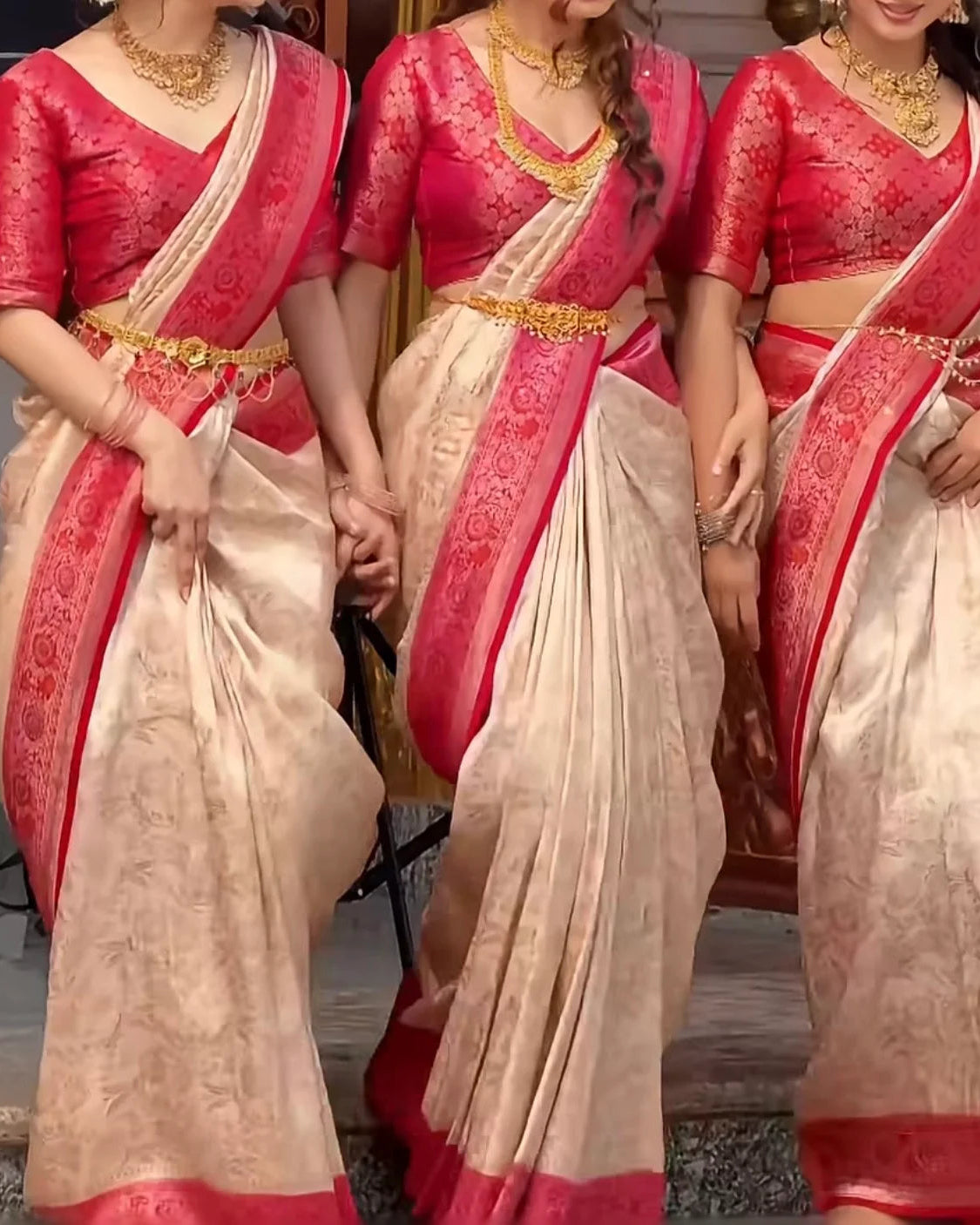 Three women wearing traditional Indian sarees with bright pinkish-red blouses and beige sarees with red borders, styled with gold jewelry.
