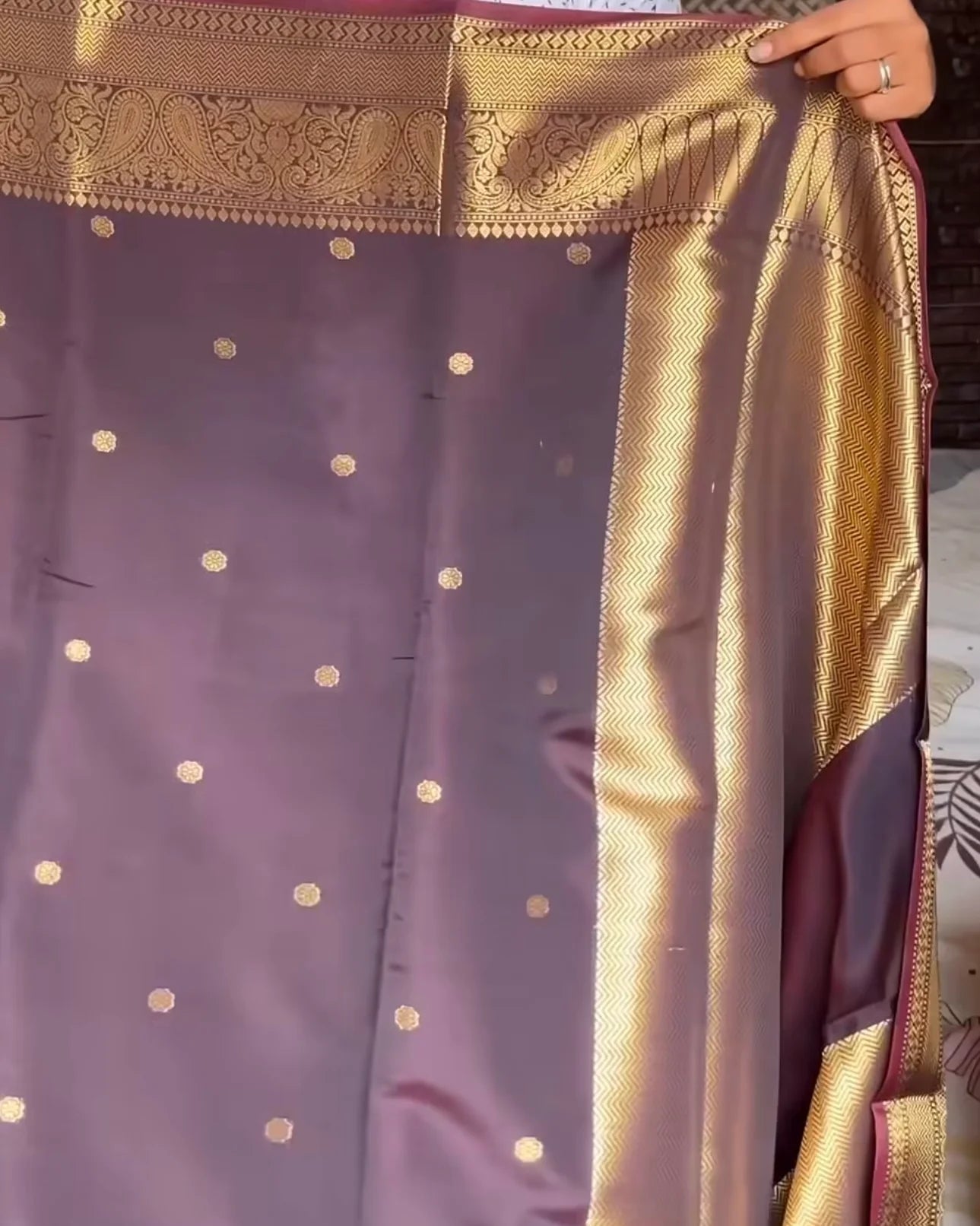 A close-up of a purple saree with gold zari motifs and a wide gold border, held up by a hand.