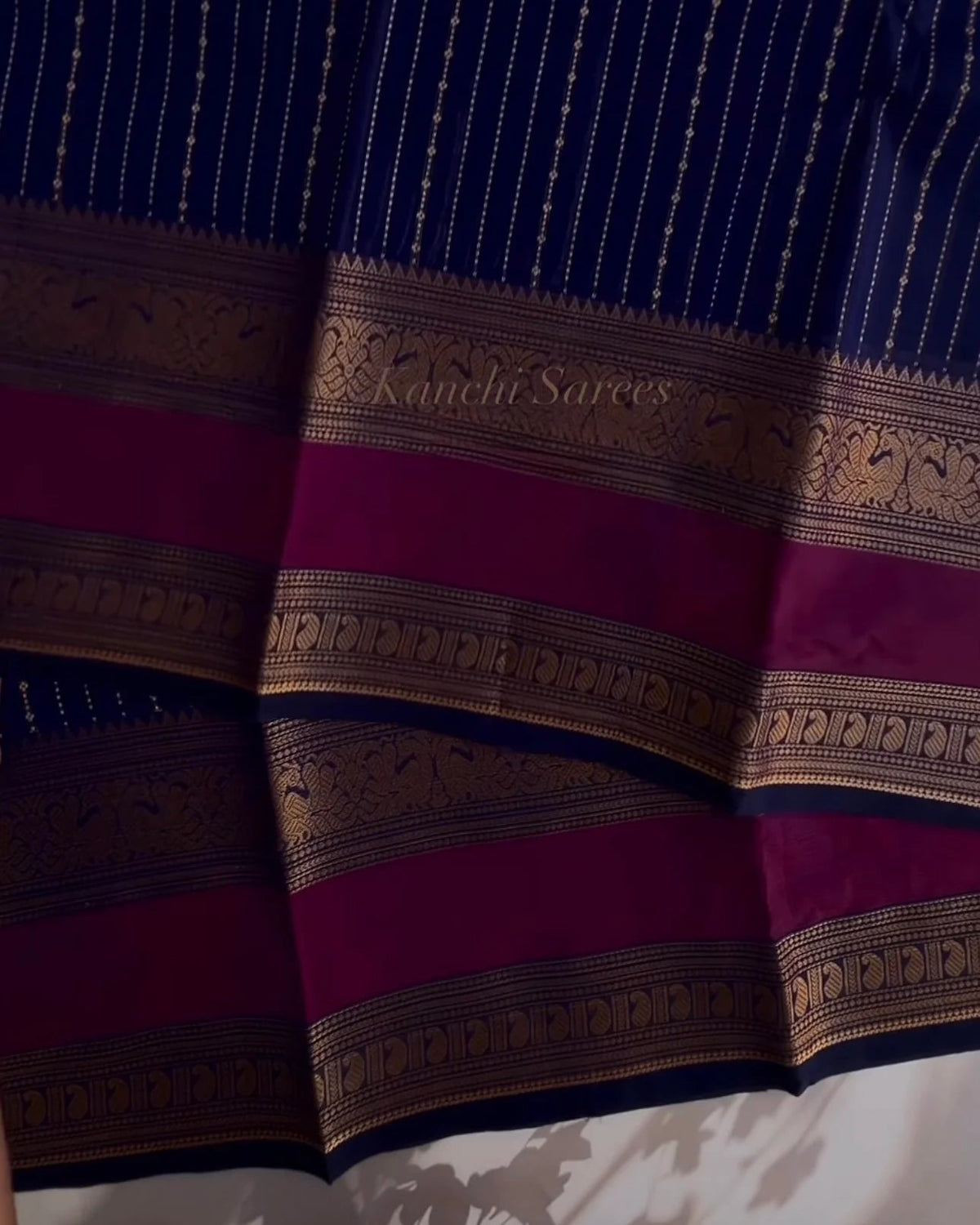 A close-up of a dark blue Kanjali saree with vertical metallic zari stripes and a wide magenta border adorned with gold zari motifs and text reading 'Kanjali Sarees'.