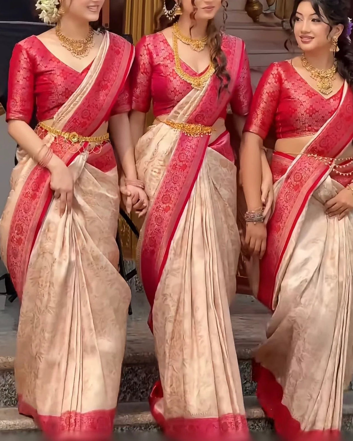 Three women wearing matching traditional Indian sarees with bright pink-red embroidered blouses and beige sarees with red borders and gold zari work.