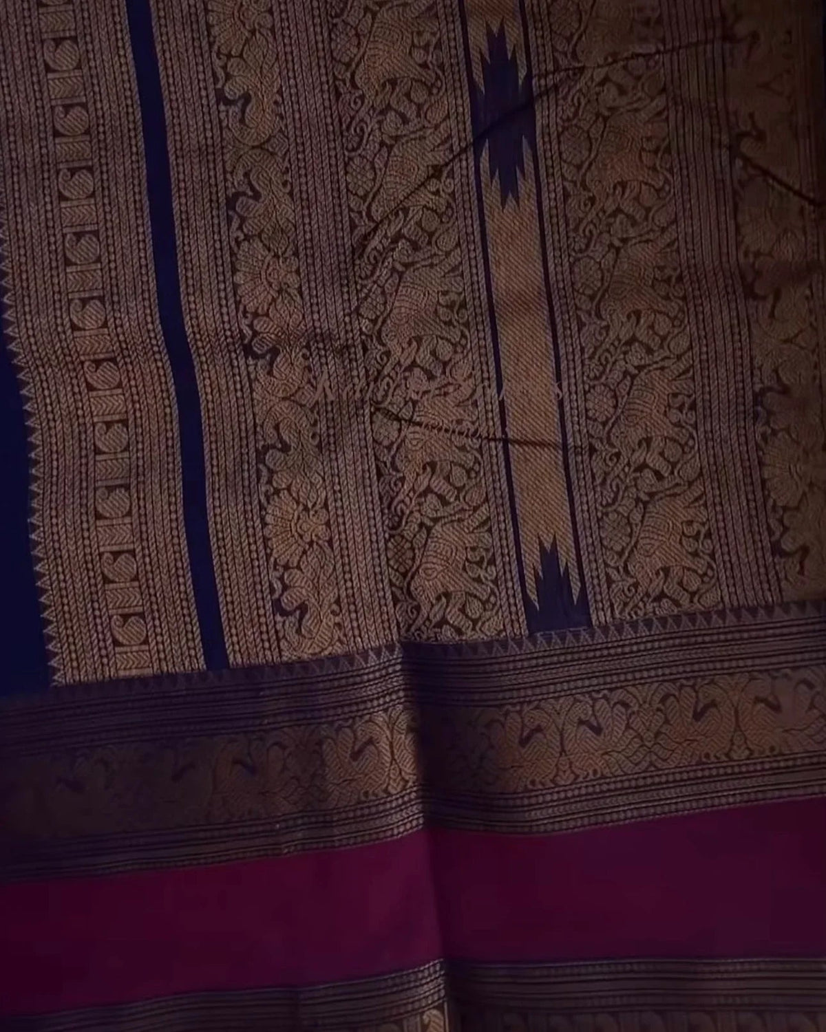 A close-up of a patterned textile, likely a sari, featuring intricate gold and navy blue woven motifs on a dark background with a prominent magenta border at the bottom.