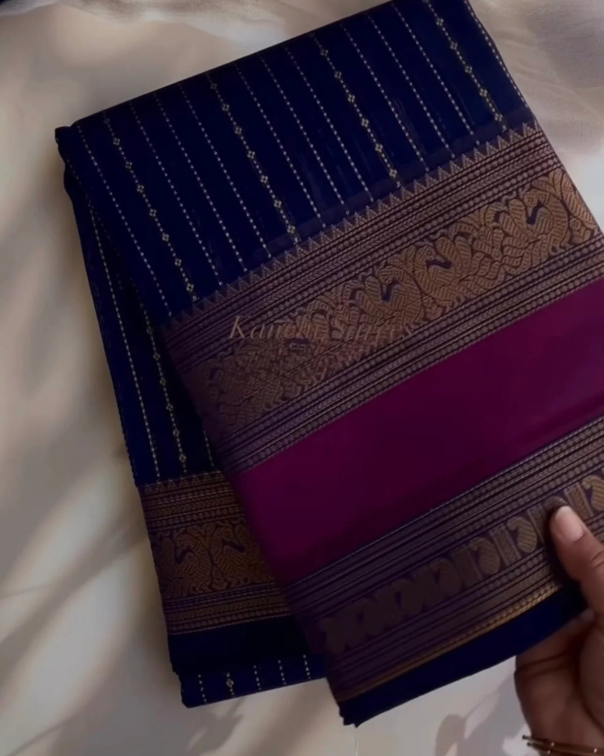 A folded dark blue and purple silk saree with intricate gold zari embroidery, held by a hand over a draped white fabric background.