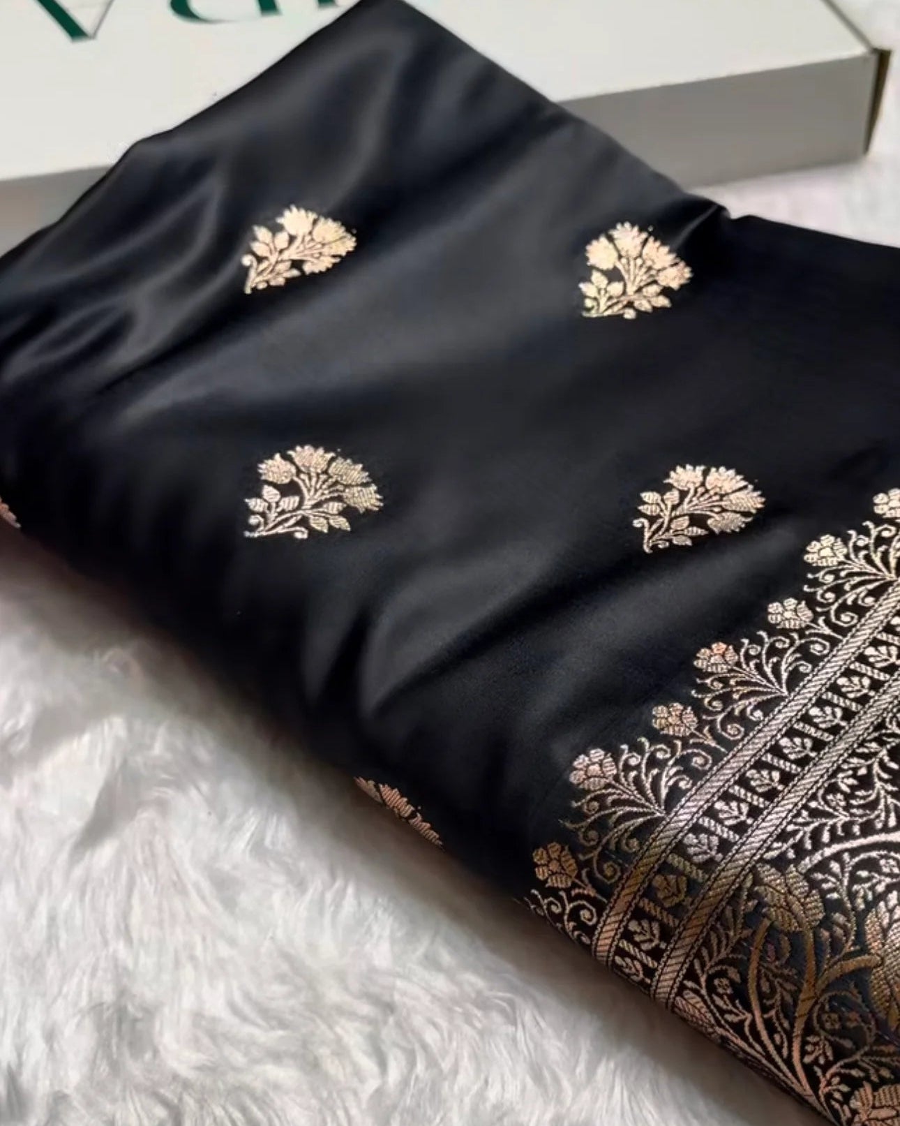 A folded black silk saree with gold floral motifs and a detailed gold border, displayed on a white faux fur surface next to its white box.