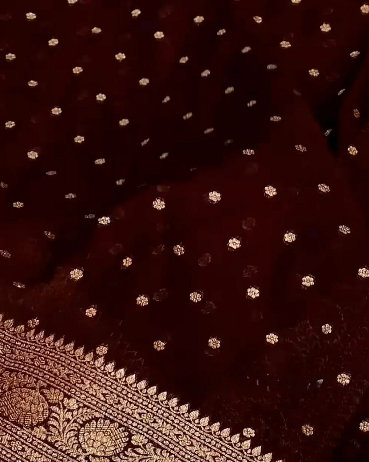 A close-up of a deep maroon or burgundy fabric with small scattered metallic gold floral motifs and a decorative gold border pattern visible along the lower edge.
