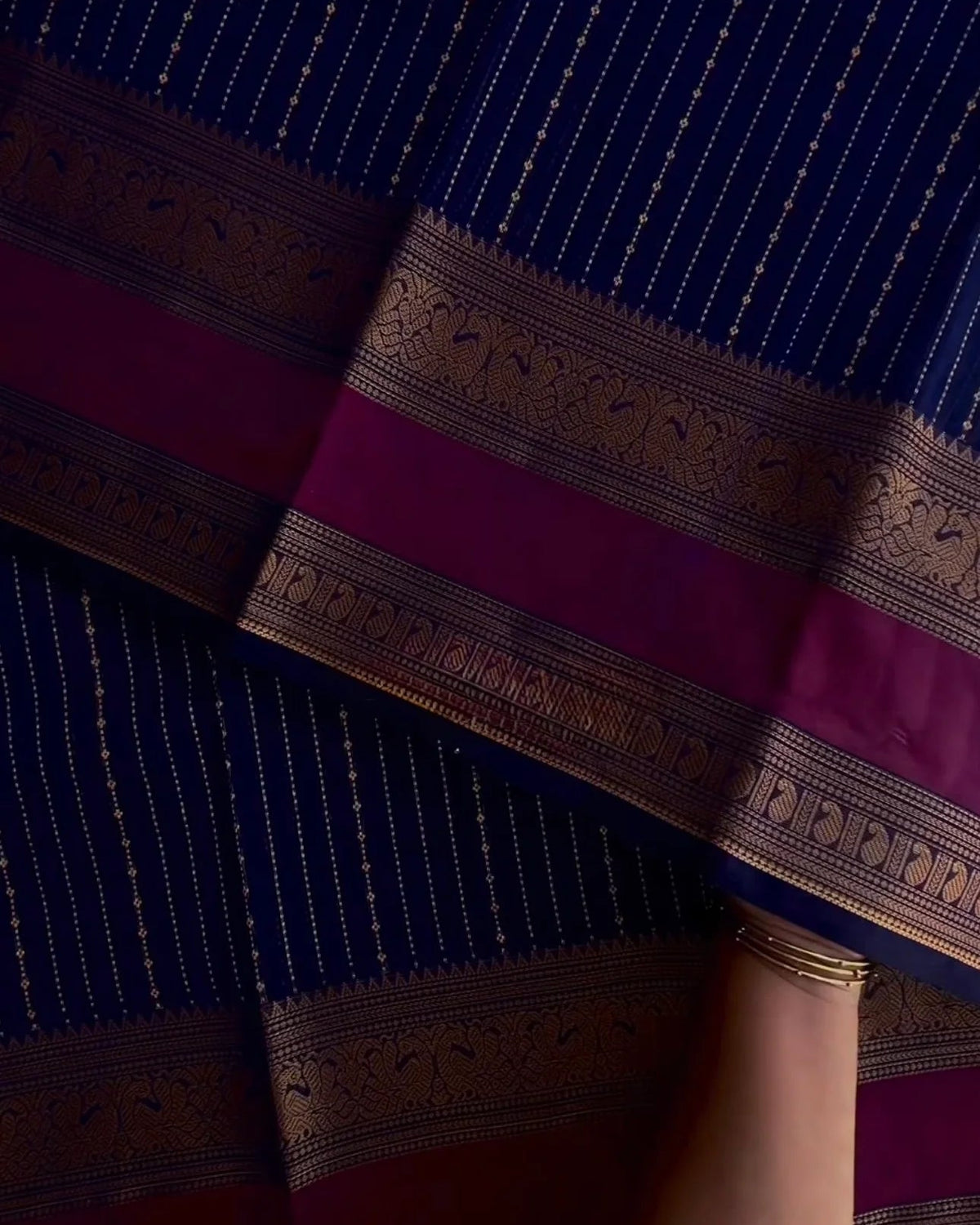 A close-up of a dark blue and magenta saree with intricate gold zari embroidery, held by a person's hand wearing a gold bangle.