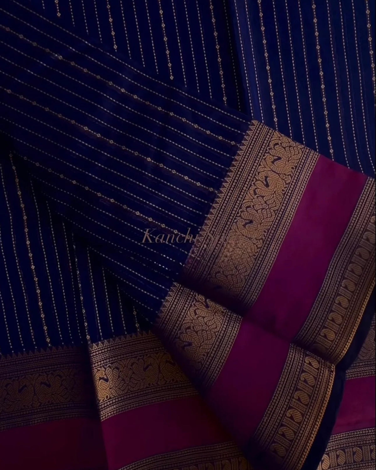 A folded dark blue silk saree with fine gold zari stripes and a rich magenta border featuring intricate gold zari motifs.