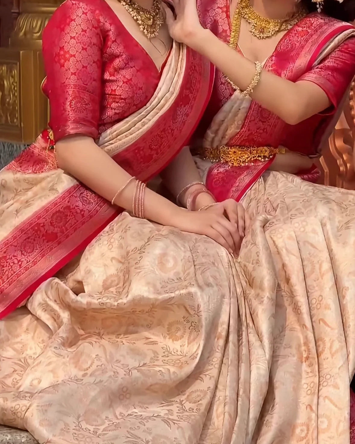 Two women seated together wearing traditional Indian sarees in rich pink and beige Banarasi silk with floral motifs, gold jewelry, and ornate carvings in the background.