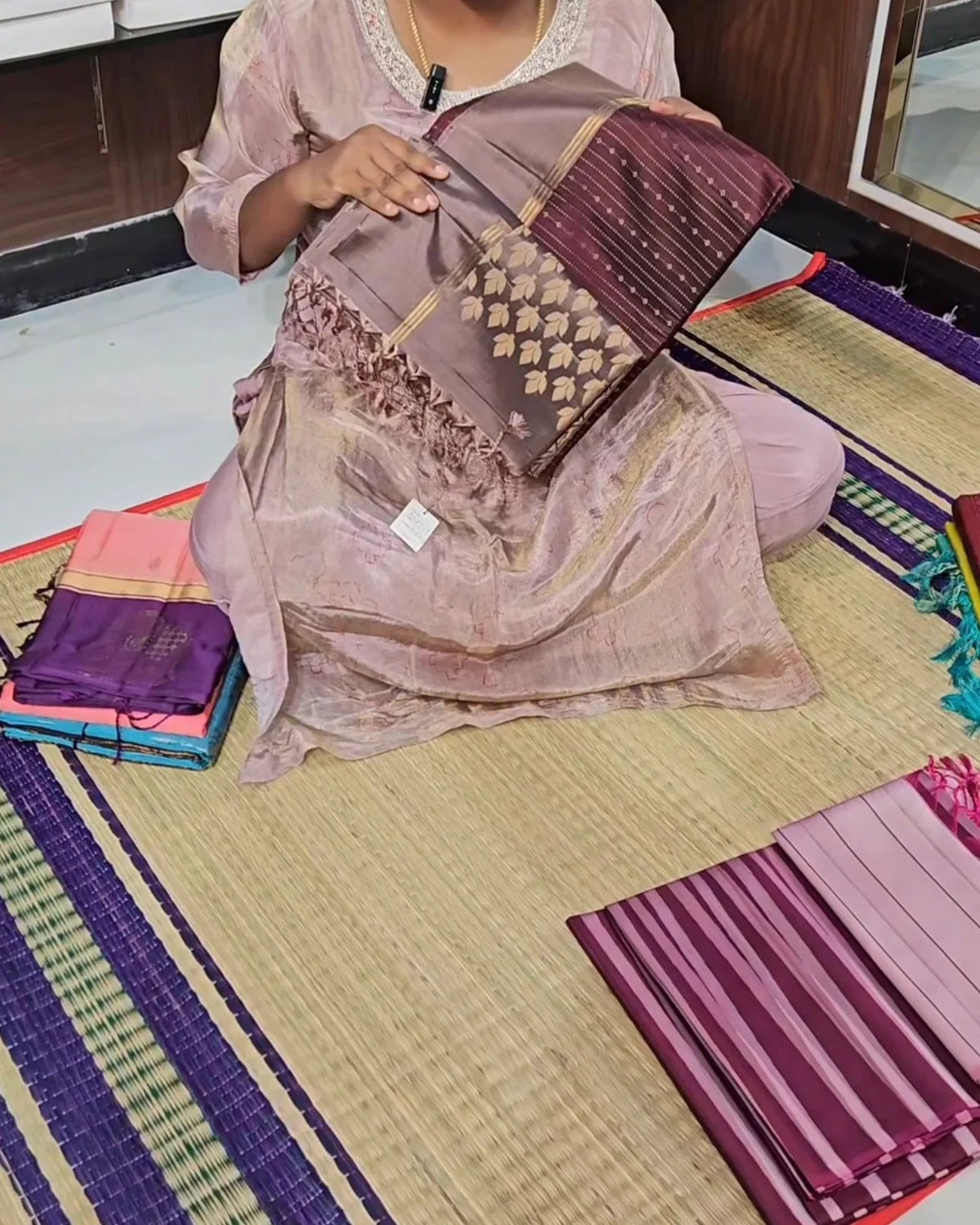 A woman sitting on a woven mat displays a folded purple and gold patterned fabric, with other folded fabrics stacked nearby on the floor.